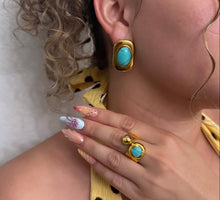 Load image into Gallery viewer, A close-up lifestyle shot of a woman wearing gold-toned jewelry, featuring a gold ring with a turquoise stone on her finger and a matching gold earring on her ear. She is wearing a yellow and black polka dot top against a white wall.