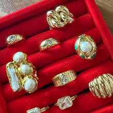 Load image into Gallery viewer, A collection of nine gold rings with white, clear, and green gemstones displayed on a red velvet ring box, set on a wooden table.