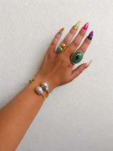 Load image into Gallery viewer, A hand with long, colorful manicured nails wearing multiple gold rings and a gold bracelet with pearl-like beads, posed against a textured off-white wall.