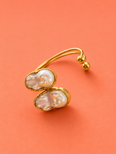 Load image into Gallery viewer, A gold-plated open cuff ring featuring two large, irregular white baroque pearls set in gold, displayed on a solid coral-orange background.