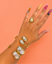 Load image into Gallery viewer, A hand and wrist wearing multiple gold rings and bracelets with large irregular white pearl-like stones, posed against a solid peach background.