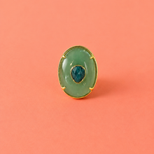Load image into Gallery viewer, An oval green cabochon ring with a gold-tone border and a small dark blue center stone, set against a solid coral pink background.