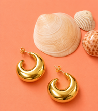 Load image into Gallery viewer, A pair of shiny gold crescent-shaped hoop earrings displayed on a peach background with decorative seashells.