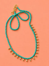 Load image into Gallery viewer, A turquoise beaded necklace with gold teardrop charms and a gold clasp, displayed on a solid coral pink background.