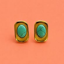 Load image into Gallery viewer, A pair of gold-toned stud earrings with oval turquoise stones, displayed on a solid coral pink background.